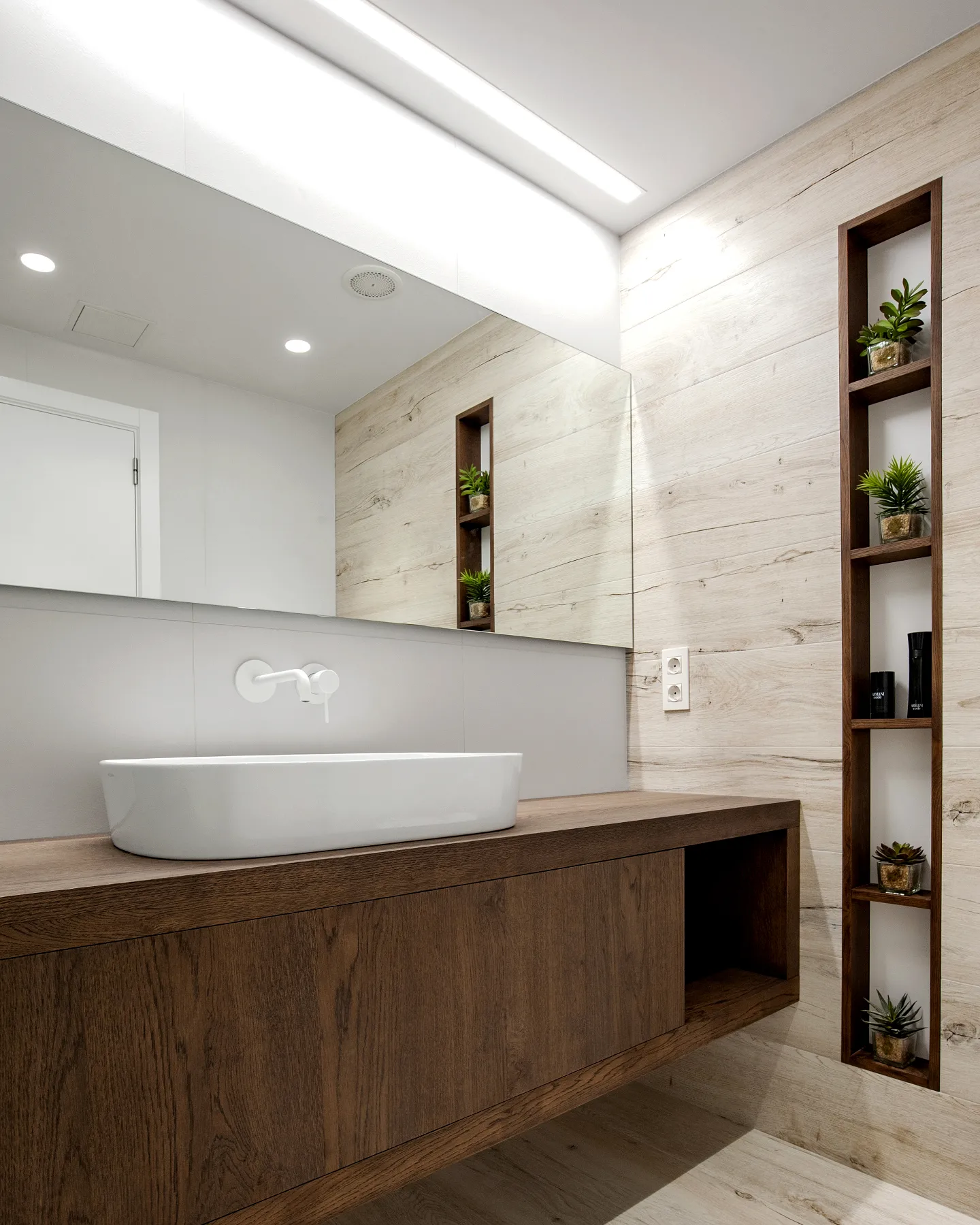 El baño de cortesía: mueble de roble oscuro, lavabo texturizado y nichos con plantas